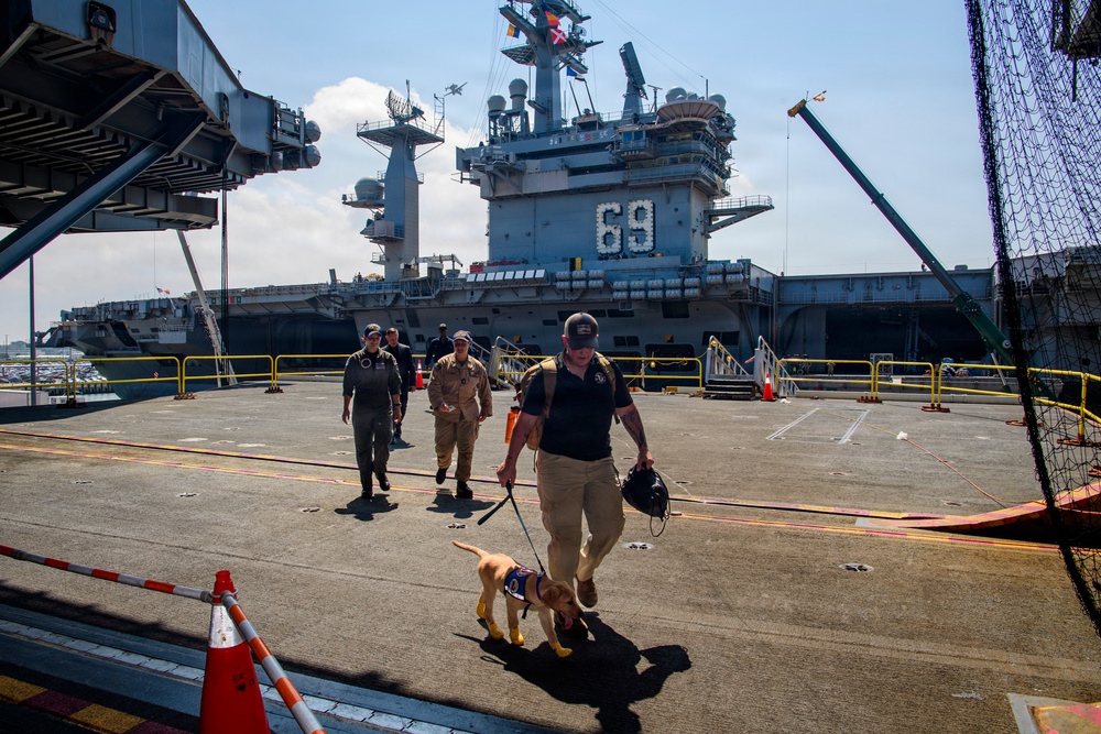 A handler leads a working dog, likely a military or security K-9, along a pier beside a large US Navy aircraft carrier bearing hull number 69. Several uniformed personnel follow behind on the dock area. The scene appears to depict a security sweep or routine inspection at a naval installation.