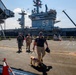 A handler leads a working dog, likely a military or security K-9, along a pier beside a large US Navy aircraft carrier bearing hull number 69. Several uniformed personnel follow behind on the dock area. The scene appears to depict a security sweep or routine inspection at a naval installation.