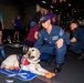 A U.S. Navy sailor in blue working uniform crouches beside a yellow Labrador Retriever therapy dog wearing a red service vest and orange protective booties, resting on a blue mat. The setting appears to be the hangar bay of a naval vessel, identifiable by the USS Nimitz (CVN-68) cap worn by the sailor. Multiple civilians and other personnel are visible in the background, suggesting a morale or community relations event.