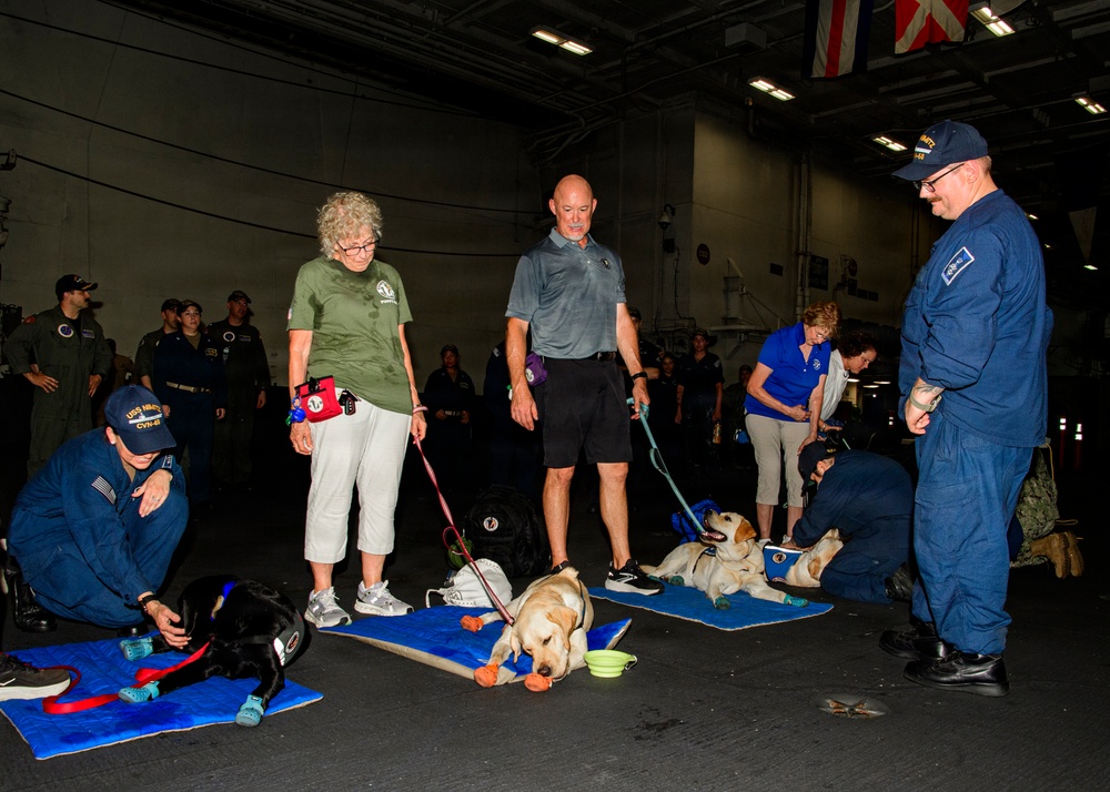 A group of people, including uniformed Navy personnel and civilian volunteers, are gathered in what appears to be a naval vessel hangar bay with therapy or service dogs resting on blue mats. The dogs, including Labrador Retrievers and a black dog, are lying on blue mats with toys and water bowls nearby. This appears to be a morale or wellness event aboard a U.S. Navy ship, likely involving a therapy dog program visit.