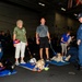 A group of people, including uniformed Navy personnel and civilian volunteers, are gathered in what appears to be a naval vessel hangar bay with therapy or service dogs resting on blue mats. The dogs, including Labrador Retrievers and a black dog, are lying on blue mats with toys and water bowls nearby. This appears to be a morale or wellness event aboard a U.S. Navy ship, likely involving a therapy dog program visit.