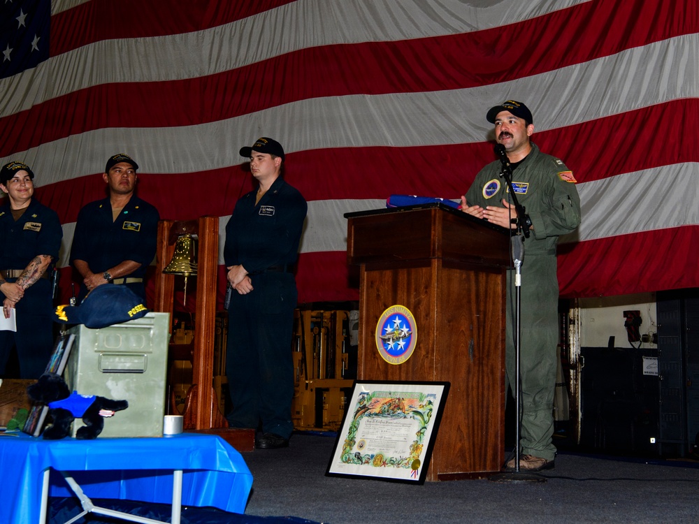 A military ceremony is taking place aboard what appears to be a U.S. Navy vessel, with an officer in a green flight suit speaking at a wooden podium bearing a naval unit insignia. A large American flag serves as a backdrop, and several sailors in dark uniforms stand at attention behind the speaker. Various ceremonial items including a ship's bell, a folded flag, and a framed certificate are visible.