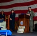 A military ceremony is taking place aboard what appears to be a U.S. Navy vessel, with an officer in a green flight suit speaking at a wooden podium bearing a naval unit insignia. A large American flag serves as a backdrop, and several sailors in dark uniforms stand at attention behind the speaker. Various ceremonial items including a ship's bell, a folded flag, and a framed certificate are visible.