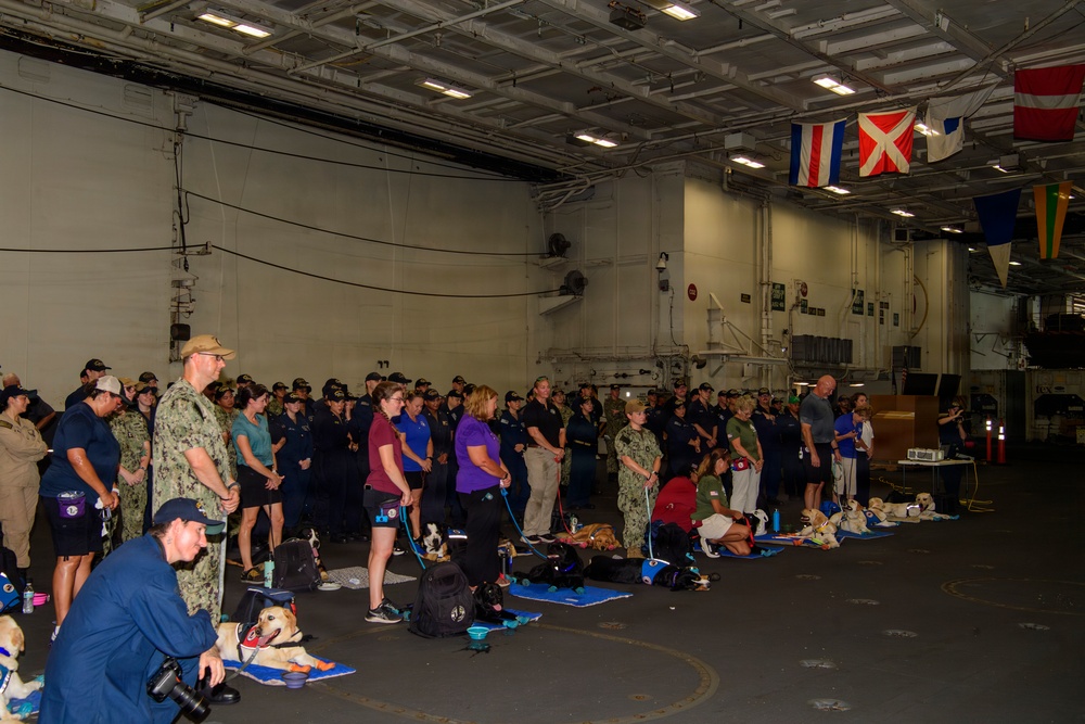 A large gathering of military personnel in Navy camouflage uniforms and civilians is taking place inside what appears to be a naval vessel's hangar bay or interior deck. Several therapy or service dogs on blue mats are present alongside their handlers, suggesting a morale or wellness event aboard the ship. Naval signal flags are visible hanging from the overhead structure in the background.