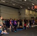 A large gathering of military personnel in Navy camouflage uniforms and civilians is taking place inside what appears to be a naval vessel's hangar bay or interior deck. Several therapy or service dogs on blue mats are present alongside their handlers, suggesting a morale or wellness event aboard the ship. Naval signal flags are visible hanging from the overhead structure in the background.