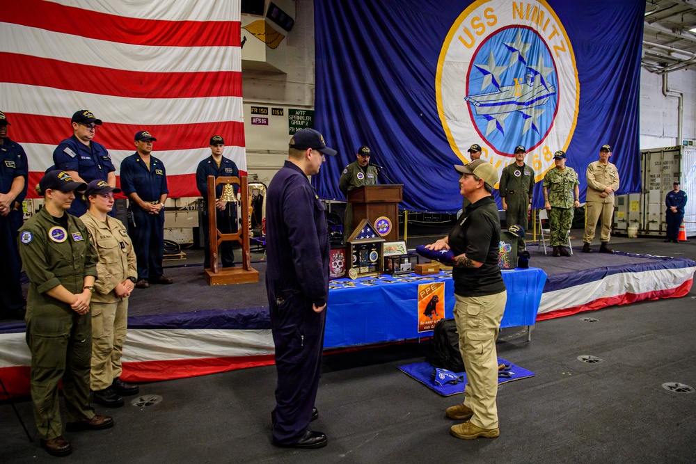 A formal military ceremony is taking place aboard the USS Nimitz (CVN-68), a U.S. Navy aircraft carrier. A civilian or off-duty personnel appears to be presenting items to a uniformed sailor in front of a display table containing awards, plaques, and memorabilia. The event is held in the ship's hangar bay, decorated with a large American flag and the USS Nimitz ship's crest banner.