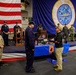 A formal military ceremony is taking place aboard the USS Nimitz (CVN-68), a U.S. Navy aircraft carrier. A civilian or off-duty personnel appears to be presenting items to a uniformed sailor in front of a display table containing awards, plaques, and memorabilia. The event is held in the ship's hangar bay, decorated with a large American flag and the USS Nimitz ship's crest banner.