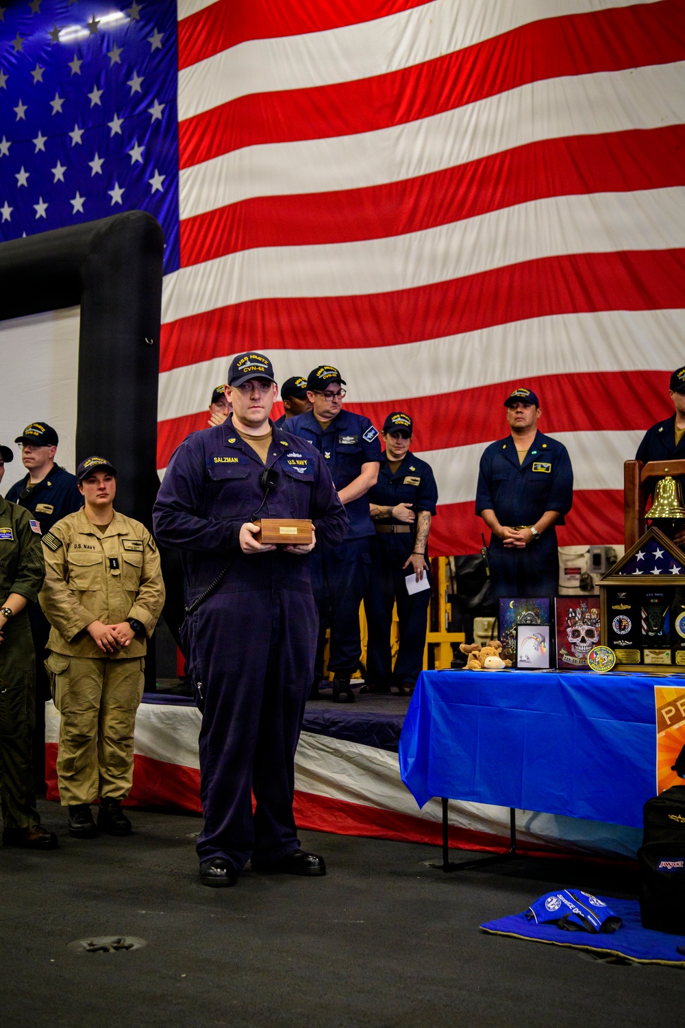 A U.S. Navy memorial ceremony is taking place in what appears to be a hangar bay, with a large American flag as a backdrop. A sailor identified by name tape as 'SALZMAN' wearing USS Nimitz CVN-68 insignia stands at the center holding a wooden box, likely an urn or memorial item. A table with memorial items including photos, plaques, a folded flag, and a ship's bell is visible to the right.