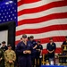 A U.S. Navy memorial ceremony is taking place in what appears to be a hangar bay, with a large American flag as a backdrop. A sailor identified by name tape as 'SALZMAN' wearing USS Nimitz CVN-68 insignia stands at the center holding a wooden box, likely an urn or memorial item. A table with memorial items including photos, plaques, a folded flag, and a ship's bell is visible to the right.