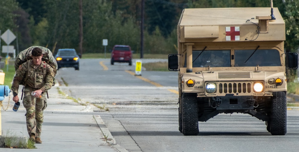 A soldier in camouflage uniform carrying a heavy rucksack walks along a road beside a tan military HMMWV (Humvee) marked with a red cross, indicating it is a medical vehicle. The scene appears to take place on a military installation or training area, with civilian vehicles visible in the background. The soldier appears to be conducting a ruck march or physical training exercise.
