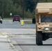 A soldier in camouflage uniform carrying a heavy rucksack walks along a road beside a tan military HMMWV (Humvee) marked with a red cross, indicating it is a medical vehicle. The scene appears to take place on a military installation or training area, with civilian vehicles visible in the background. The soldier appears to be conducting a ruck march or physical training exercise.