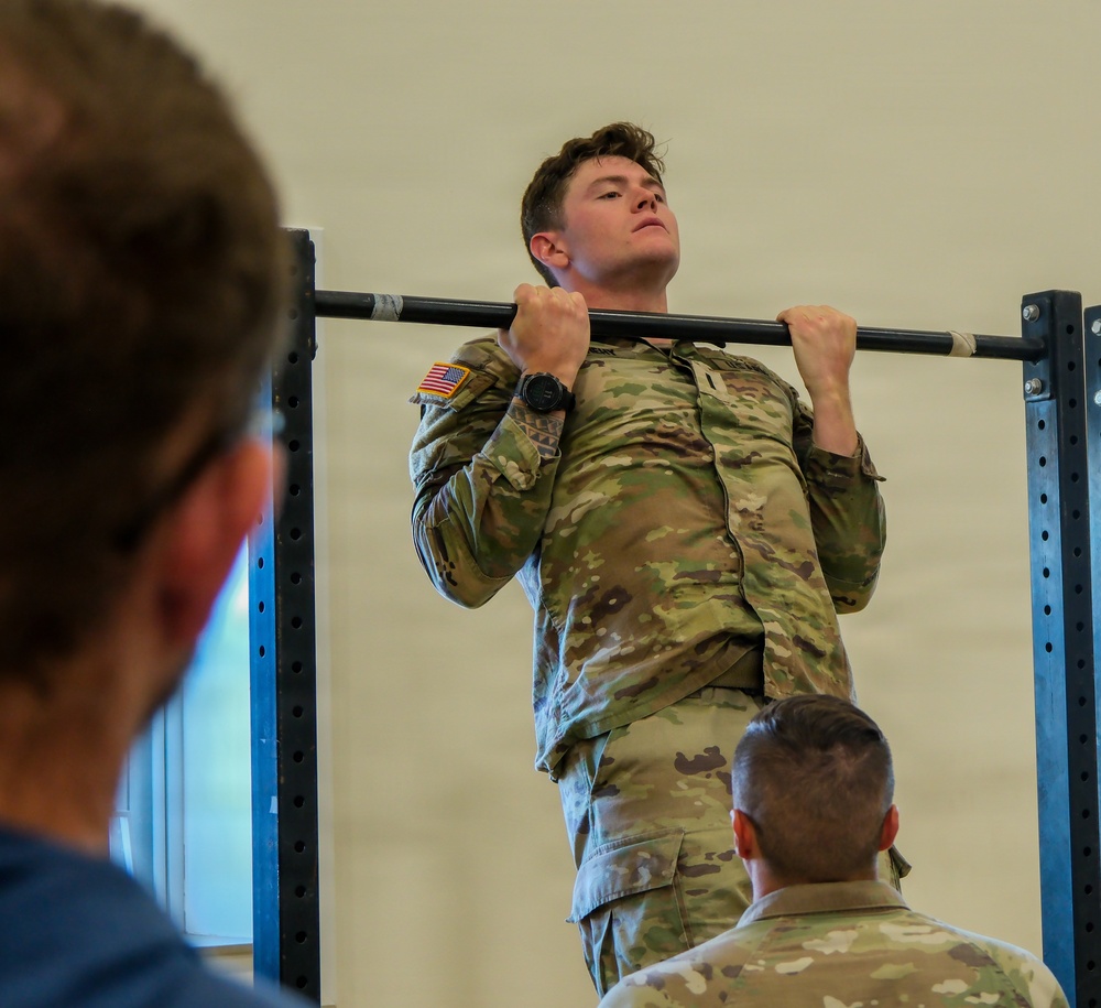 A U.S. Army soldier in OCP (Operational Camouflage Pattern) uniform is performing pull-ups on a bar mounted to a blue power rack inside what appears to be a gymnasium or fitness facility. The soldier wears a U.S. flag patch on the left shoulder and a tactical watch on the left wrist. At least two other individuals are visible in the foreground observing, suggesting this may be a fitness test or evaluation.