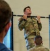 A U.S. Army soldier in OCP (Operational Camouflage Pattern) uniform is performing pull-ups on a bar mounted to a blue power rack inside what appears to be a gymnasium or fitness facility. The soldier wears a U.S. flag patch on the left shoulder and a tactical watch on the left wrist. At least two other individuals are visible in the foreground observing, suggesting this may be a fitness test or evaluation.