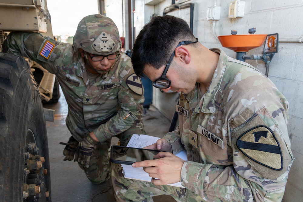 Two U.S. Army soldiers in Operational Camouflage Pattern (OCP) uniforms are conducting a vehicle maintenance inspection near a large military truck tire. The soldier in the foreground, identified by the name tape 'PERTH' and wearing a 1st Cavalry Division patch, is reviewing documents on a tablet and paper forms. The second soldier, identified as 'TOM', is wearing a helmet and examining the vehicle wheel area.