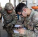 Two U.S. Army soldiers in Operational Camouflage Pattern (OCP) uniforms are conducting a vehicle maintenance inspection near a large military truck tire. The soldier in the foreground, identified by the name tape 'PERTH' and wearing a 1st Cavalry Division patch, is reviewing documents on a tablet and paper forms. The second soldier, identified as 'TOM', is wearing a helmet and examining the vehicle wheel area.