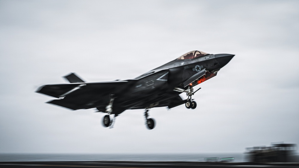 A U.S. military F-35B stealth fighter jet is captured mid-takeoff or short landing from what appears to be a naval vessel, likely an aircraft carrier or amphibious assault ship. The aircraft displays U.S. military markings including a star-and-bar insignia and has its landing gear deployed. The image was taken with motion blur techniques emphasizing speed and dynamic movement.