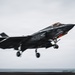 A U.S. military F-35B stealth fighter jet is captured mid-takeoff or short landing from what appears to be a naval vessel, likely an aircraft carrier or amphibious assault ship. The aircraft displays U.S. military markings including a star-and-bar insignia and has its landing gear deployed. The image was taken with motion blur techniques emphasizing speed and dynamic movement.
