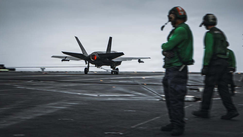 A fifth-generation stealth fighter jet, identifiable as an F-35C, is landing on an aircraft carrier flight deck with its tailhook engaged on the arresting wire. Two flight deck crew members wearing green jerseys and helmets observe the arrested landing from the right side of the frame. The overcast sky and wet deck surface suggest operations in challenging weather conditions.
