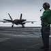 A fifth-generation stealth fighter jet, identifiable as an F-35C, is landing on an aircraft carrier flight deck with its tailhook engaged on the arresting wire. Two flight deck crew members wearing green jerseys and helmets observe the arrested landing from the right side of the frame. The overcast sky and wet deck surface suggest operations in challenging weather conditions.