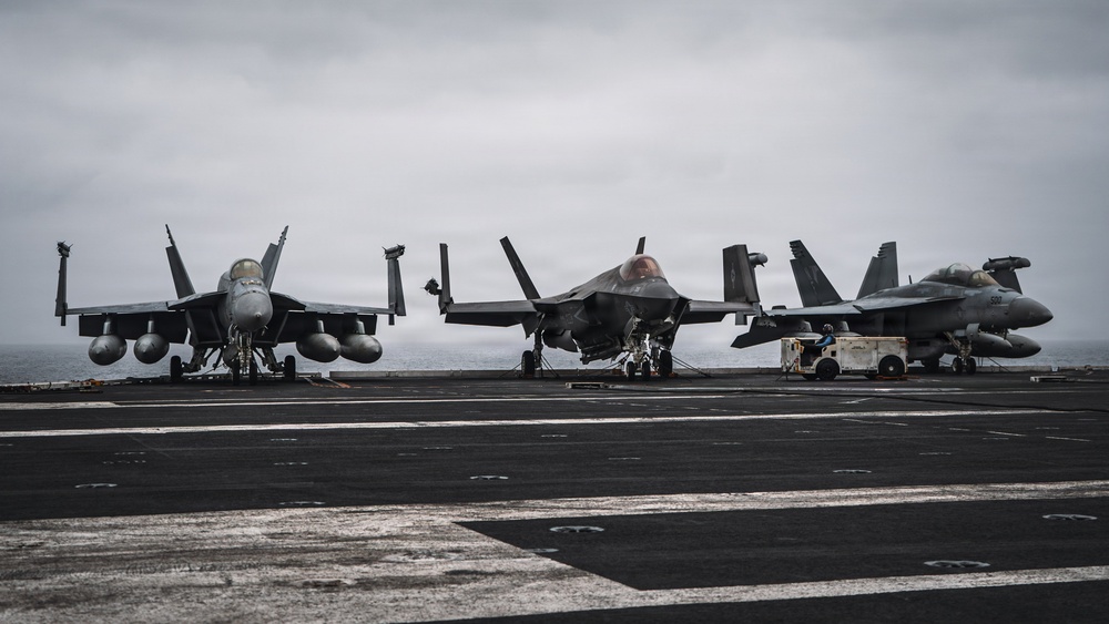 Three military jet aircraft are parked on the flight deck of a naval aircraft carrier at sea under overcast skies. The aircraft include what appear to be two F/A-18 Super Hornets flanking a center F-35C Lightning II, representing a mix of legacy and fifth-generation carrier-based fighters. Ground crew personnel and support equipment are visible on the deck.