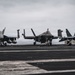 Three military jet aircraft are parked on the flight deck of a naval aircraft carrier at sea under overcast skies. The aircraft include what appear to be two F/A-18 Super Hornets flanking a center F-35C Lightning II, representing a mix of legacy and fifth-generation carrier-based fighters. Ground crew personnel and support equipment are visible on the deck.