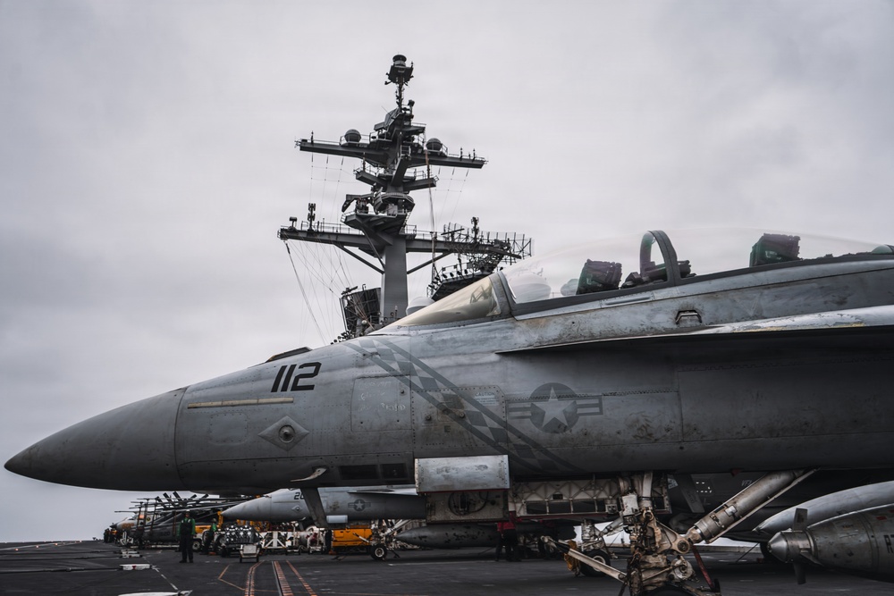 A U.S. Navy carrier-based fighter jet, marked with the number '112' and a U.S. military roundel, is parked on the flight deck of an aircraft carrier. The carrier's island superstructure, featuring radar arrays and communication equipment, is prominently visible in the background. The scene appears to be during overcast conditions with deck crew and support equipment visible nearby.
