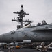 A U.S. Navy carrier-based fighter jet, marked with the number '112' and a U.S. military roundel, is parked on the flight deck of an aircraft carrier. The carrier's island superstructure, featuring radar arrays and communication equipment, is prominently visible in the background. The scene appears to be during overcast conditions with deck crew and support equipment visible nearby.