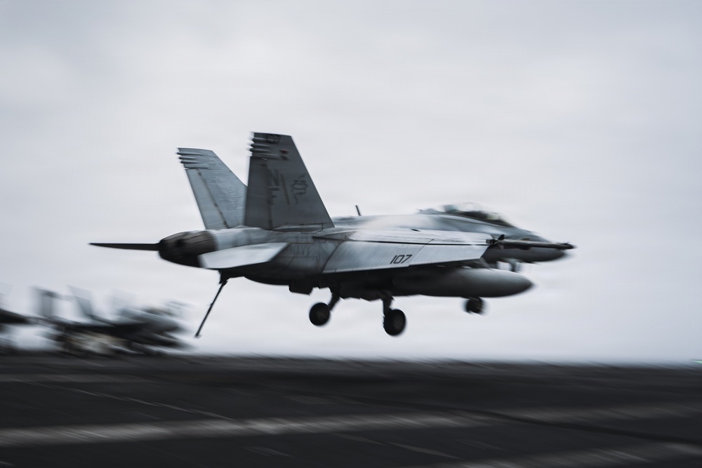 A U.S. Navy F/A-18 Super Hornet (bureau number 107) is captured in motion during a carrier landing or takeoff on what appears to be an aircraft carrier flight deck. The image features intentional motion blur conveying high speed and dynamic action. The overcast sky and dark flight deck surface are characteristic of open-ocean carrier operations.