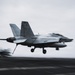 A U.S. Navy F/A-18 Super Hornet (bureau number 107) is captured in motion during a carrier landing or takeoff on what appears to be an aircraft carrier flight deck. The image features intentional motion blur conveying high speed and dynamic action. The overcast sky and dark flight deck surface are characteristic of open-ocean carrier operations.