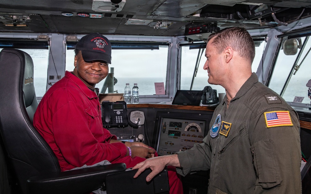 Two individuals are photographed on what appears to be the bridge of a naval vessel. One person in a red jumpsuit and black cap is seated at the helm controls, while a military officer in an olive flight suit with U.S. flag and unit patches stands beside them. The setting includes navigation equipment, communication devices, and ocean visible through the bridge windows.