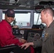 Two individuals are photographed on what appears to be the bridge of a naval vessel. One person in a red jumpsuit and black cap is seated at the helm controls, while a military officer in an olive flight suit with U.S. flag and unit patches stands beside them. The setting includes navigation equipment, communication devices, and ocean visible through the bridge windows.