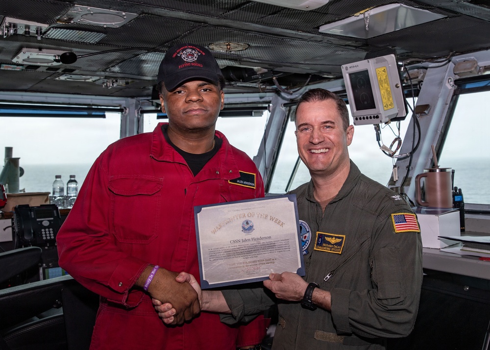 Two military personnel are posing for a photo aboard what appears to be a naval vessel's bridge or command area. They are jointly holding a certificate titled 'Sailor of the Week' awarded to CSSN Jalen Henderson, while shaking hands. The man on the right, identified by his name tag as Nicholas Dean with the title 'Commanding Officer,' is presenting the award.