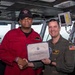 Two military personnel are posing for a photo aboard what appears to be a naval vessel's bridge or command area. They are jointly holding a certificate titled 'Sailor of the Week' awarded to CSSN Jalen Henderson, while shaking hands. The man on the right, identified by his name tag as Nicholas Dean with the title 'Commanding Officer,' is presenting the award.