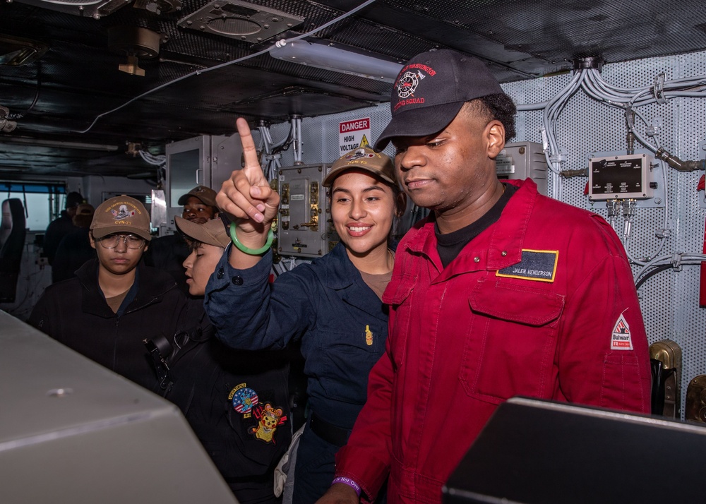 A group of U.S. Navy personnel are gathered inside what appears to be a ship's interior compartment, likely aboard a naval vessel. A sailor in a navy blue uniform is pointing upward while standing next to a crew member named 'Jalen Henderson' wearing a red coverall uniform. The setting includes visible safety signage, electrical panels, and ship infrastructure.