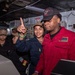 A group of U.S. Navy personnel are gathered inside what appears to be a ship's interior compartment, likely aboard a naval vessel. A sailor in a navy blue uniform is pointing upward while standing next to a crew member named 'Jalen Henderson' wearing a red coverall uniform. The setting includes visible safety signage, electrical panels, and ship infrastructure.