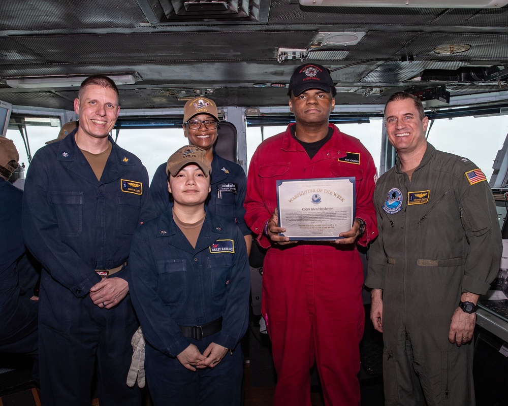 A group of five U.S. Navy personnel pose together on what appears to be a ship's bridge or command area. The central figure, wearing a red flight deck jumpsuit, is holding a 'Warfighter of the Week' certificate awarded to 'CSSN Jalen Henderson'. The group includes personnel in standard Navy working uniforms and one individual in an olive/green flight suit.