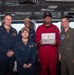A group of five U.S. Navy personnel pose together on what appears to be a ship's bridge or command area. The central figure, wearing a red flight deck jumpsuit, is holding a 'Warfighter of the Week' certificate awarded to 'CSSN Jalen Henderson'. The group includes personnel in standard Navy working uniforms and one individual in an olive/green flight suit.