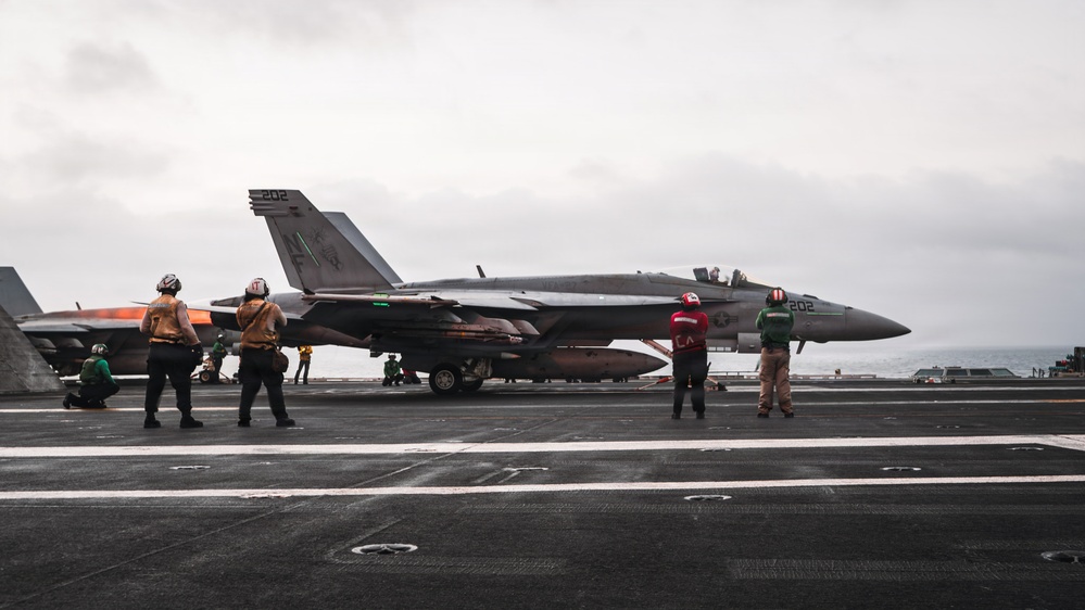 A carrier-based military jet aircraft marked '202' is positioned on the flight deck of a naval aircraft carrier, surrounded by deck crew in colored jerseys. Personnel in yellow/brown, red, and green jerseys are visible performing pre-flight or deck operations. The overcast sky and visible ocean horizon confirm an open-sea carrier operations environment.