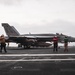 A carrier-based military jet aircraft marked '202' is positioned on the flight deck of a naval aircraft carrier, surrounded by deck crew in colored jerseys. Personnel in yellow/brown, red, and green jerseys are visible performing pre-flight or deck operations. The overcast sky and visible ocean horizon confirm an open-sea carrier operations environment.