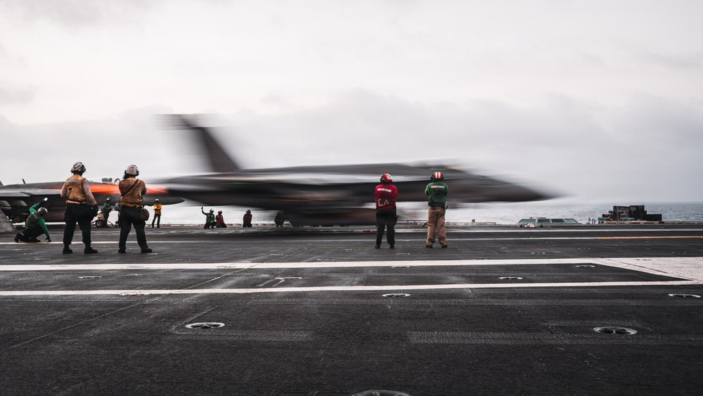 A military jet aircraft is captured mid-launch from the deck of an aircraft carrier, with significant motion blur indicating high speed. Several flight deck crew members in color-coded jerseys are visible observing or directing the launch operation. The overcast sky and open ocean are visible in the background.