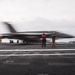 A military jet aircraft is captured mid-launch from the deck of an aircraft carrier, with significant motion blur indicating high speed. Several flight deck crew members in color-coded jerseys are visible observing or directing the launch operation. The overcast sky and open ocean are visible in the background.
