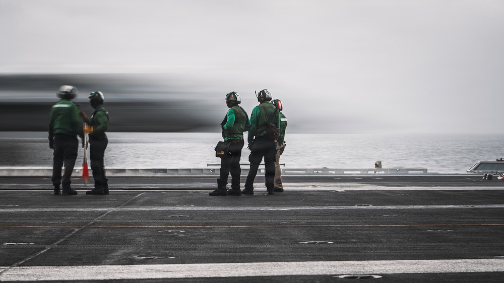 Four personnel in green jerseys and helmets stand on what appears to be an aircraft carrier flight deck, watching a fast-moving aircraft (visible as a motion-blurred streak) launch or pass by. The scene is set over open ocean under overcast skies. The green jerseys indicate these are likely aircraft handling or catapult/arresting gear crew members.