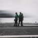 Four personnel in green jerseys and helmets stand on what appears to be an aircraft carrier flight deck, watching a fast-moving aircraft (visible as a motion-blurred streak) launch or pass by. The scene is set over open ocean under overcast skies. The green jerseys indicate these are likely aircraft handling or catapult/arresting gear crew members.