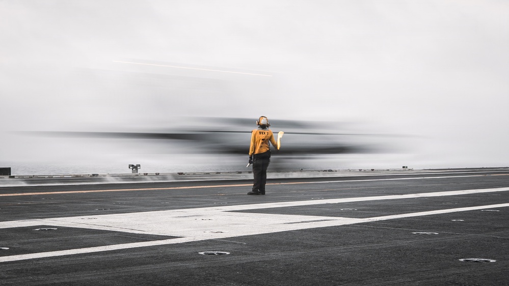 A flight deck crew member wearing a yellow jersey labeled 'FLY 2' stands on an aircraft carrier flight deck as a fast-moving aircraft passes by, captured with motion blur. The person is facing away from the camera, holding what appears to be signal wands, watching the aircraft. The image uses selective color, keeping the crew member in color while the surroundings remain largely desaturated.