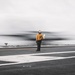A flight deck crew member wearing a yellow jersey labeled 'FLY 2' stands on an aircraft carrier flight deck as a fast-moving aircraft passes by, captured with motion blur. The person is facing away from the camera, holding what appears to be signal wands, watching the aircraft. The image uses selective color, keeping the crew member in color while the surroundings remain largely desaturated.