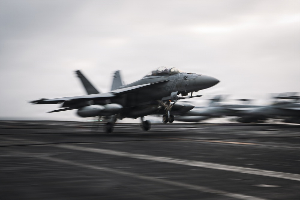 A U.S. Navy F/A-18 Super Hornet (bureau number 112) is captured launching or landing on an aircraft carrier flight deck. The image is taken with a panning technique, creating motion blur in the background while keeping the aircraft relatively sharp. The overcast sky and dark non-skid flight deck surface are characteristic of carrier operations at sea.
