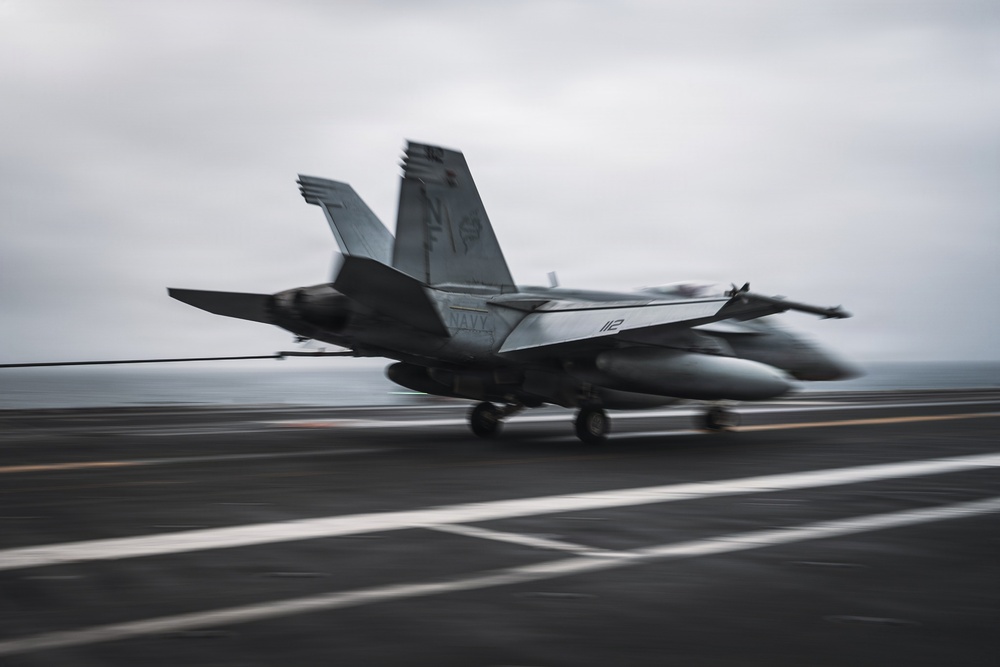 A U.S. Navy jet aircraft, marked with the number '112' and 'NAVY' on its fuselage, is captured mid-landing or arrested landing on what appears to be an aircraft carrier flight deck. The motion blur effect conveys the high speed of the aircraft as it engages the arresting wire visible stretching across the deck. The overcast sky and open ocean horizon in the background confirm an at-sea carrier operation.