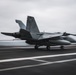 A U.S. Navy jet aircraft, marked with the number '112' and 'NAVY' on its fuselage, is captured mid-landing or arrested landing on what appears to be an aircraft carrier flight deck. The motion blur effect conveys the high speed of the aircraft as it engages the arresting wire visible stretching across the deck. The overcast sky and open ocean horizon in the background confirm an at-sea carrier operation.