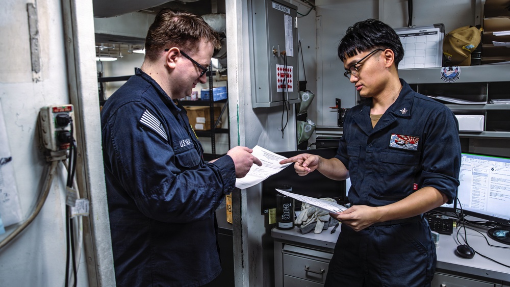 Two U.S. Navy personnel in dark blue utility uniforms are reviewing documents together in what appears to be a shipboard workspace or supply office. One sailor is pointing to a specific section of a document held by the other, suggesting a briefing, inspection, or administrative review. The setting includes shelving, a computer workstation, and various naval equipment consistent with an interior ship compartment.