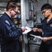 Two U.S. Navy personnel in dark blue utility uniforms are reviewing documents together in what appears to be a shipboard workspace or supply office. One sailor is pointing to a specific section of a document held by the other, suggesting a briefing, inspection, or administrative review. The setting includes shelving, a computer workstation, and various naval equipment consistent with an interior ship compartment.